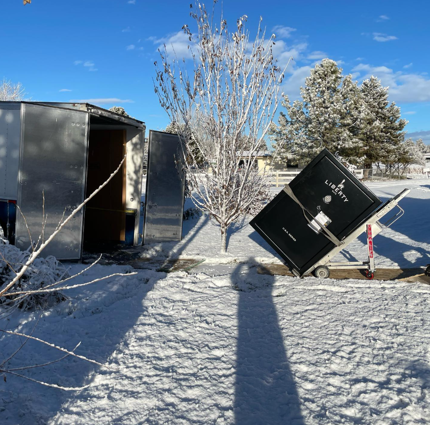 Liberty safe tilted on a hand truck beside an open trailer in a snowy residential yard.